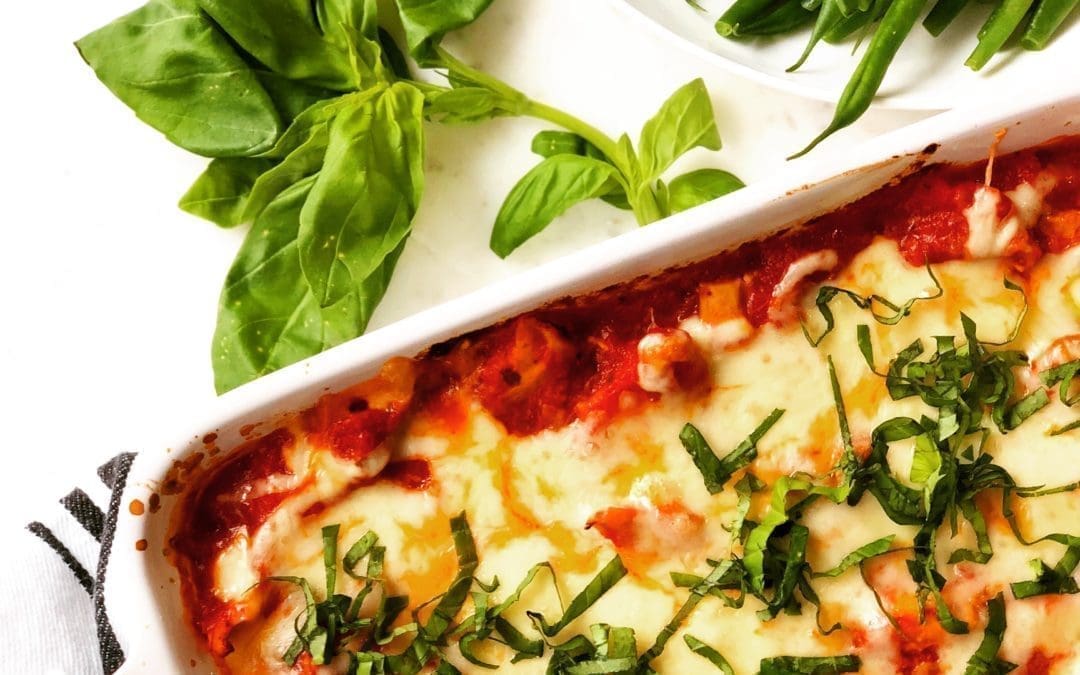 A white baking dish of Healthier Chicken Parmesan Casserole is seen from above, surrounded by fresh basil and a bowl of steamed green beans.