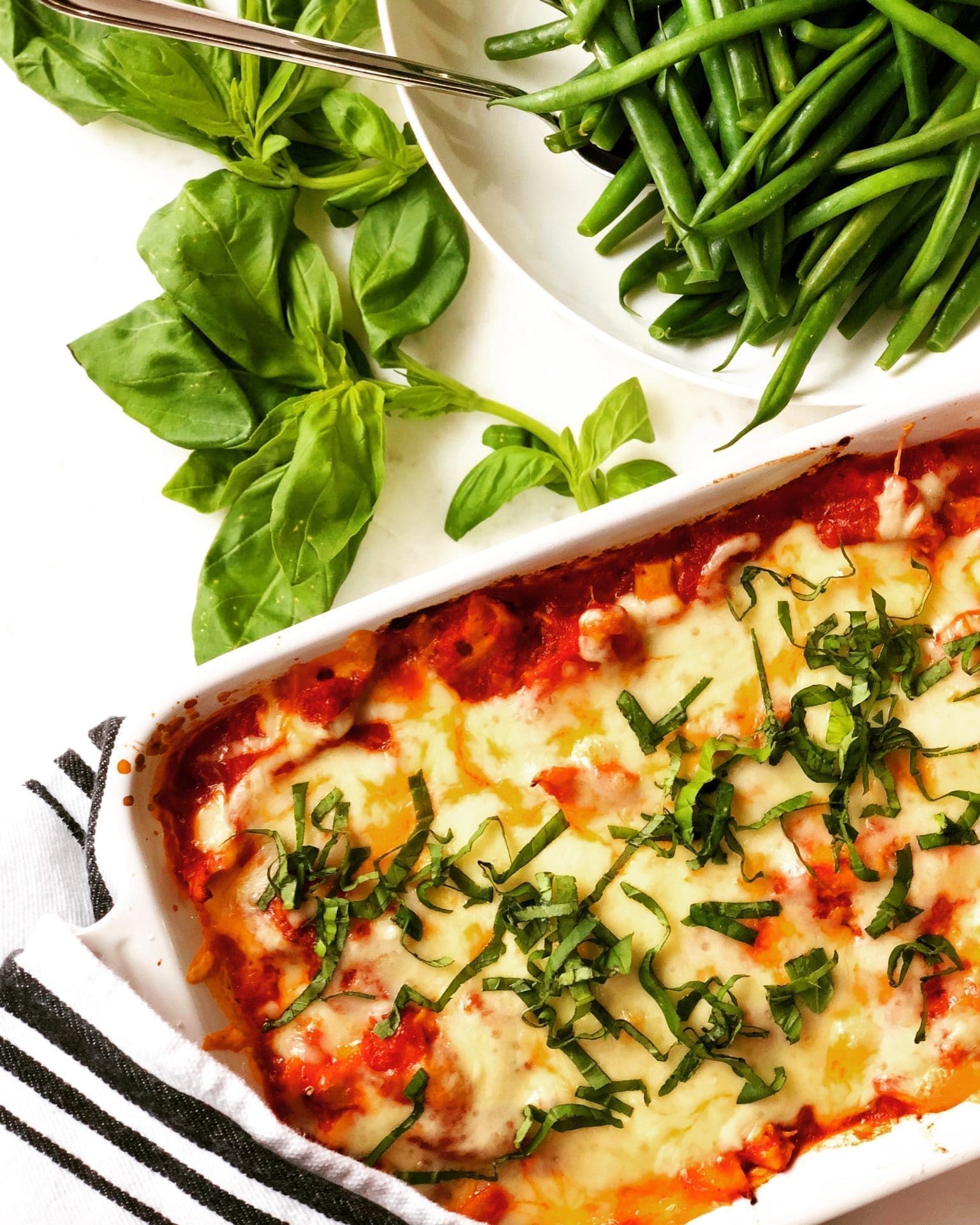 A white baking dish of Healthier Chicken Parmesan Casserole is seen from above, surrounded by fresh basil and a bowl of steamed green beans.