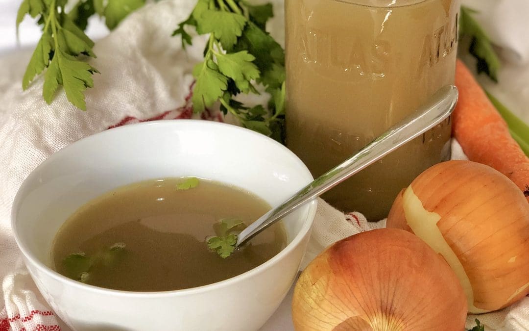 A bowl of homemade golden chicken stock sits on a white table, surrounded by fresh vegetables and herbs