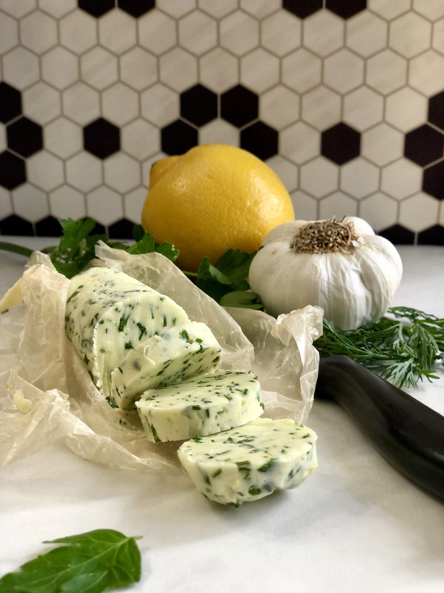 0E259917-D6BF-4FD2-A62D-F178112FBB63 A log of chilled herb and garlic butter is sliced into rounds and seen on a counter surrounded by lemon, garlic and herbs.