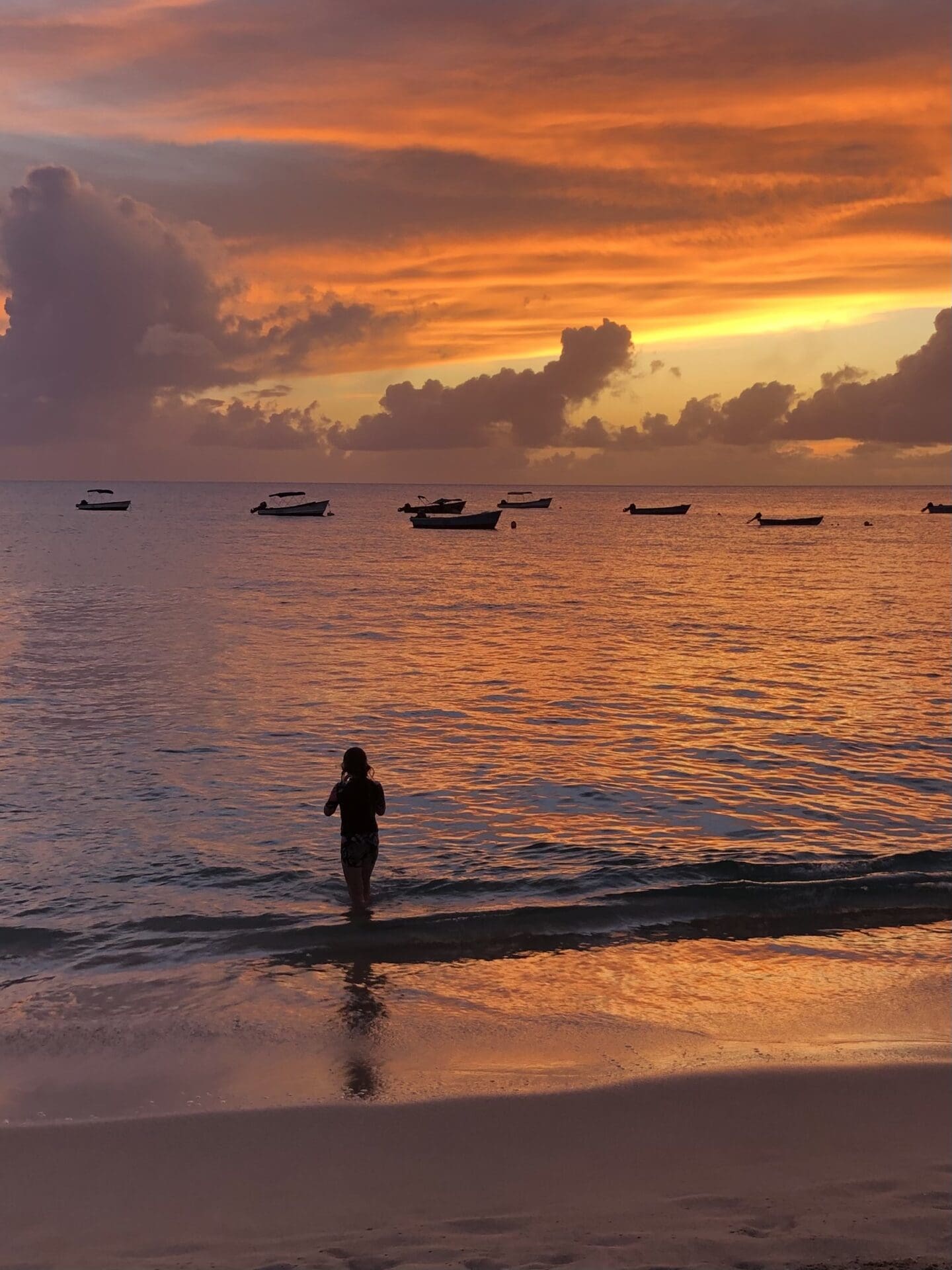 Stunning sunset on a Barbados beach with boats bobbing in the background and woman emerging from a swim in the ocean