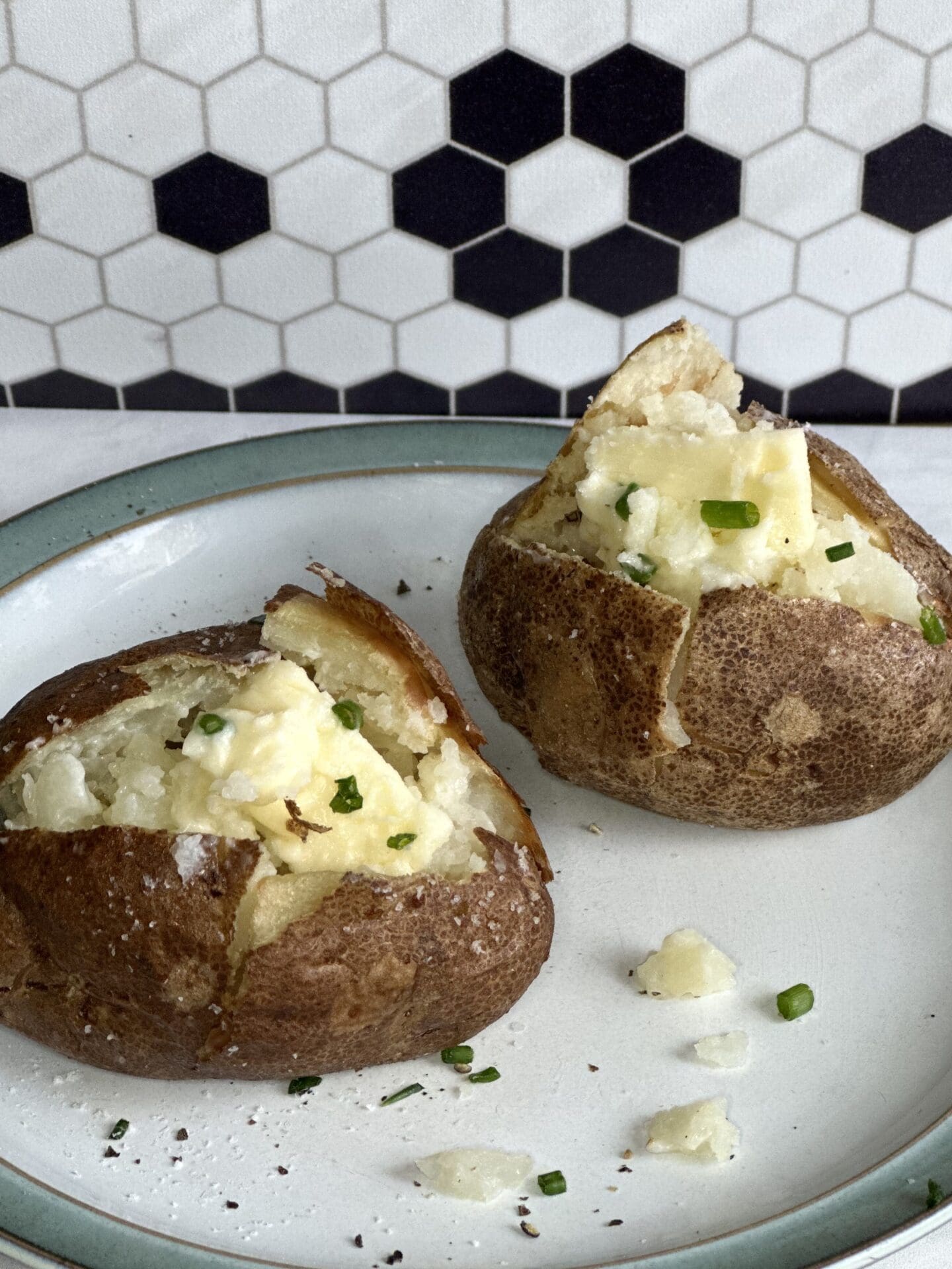 45045EBC-BBBA-470F-AB08-6C17164418E1 Plate of 2 perfect baked potatoes, split open to reveal a fluffy interior. Potatoes are topped with pats of butter and sprinkles of chives. Plate is sitting on a white marble counter top with a black and white mosaic backsplash seen in the background.
