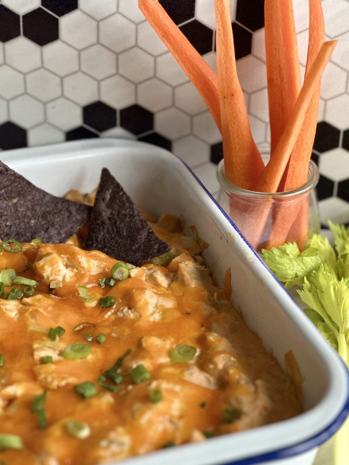 9FFF5575-2C1F-428F-B02B-AEE24211FA12 White enamel pan of Hot and Spicy Buffalo Chicken Dip with a topping of melted cheddar and green onions. Seen on a counter top with vegetables and blue corn chips for dipping.