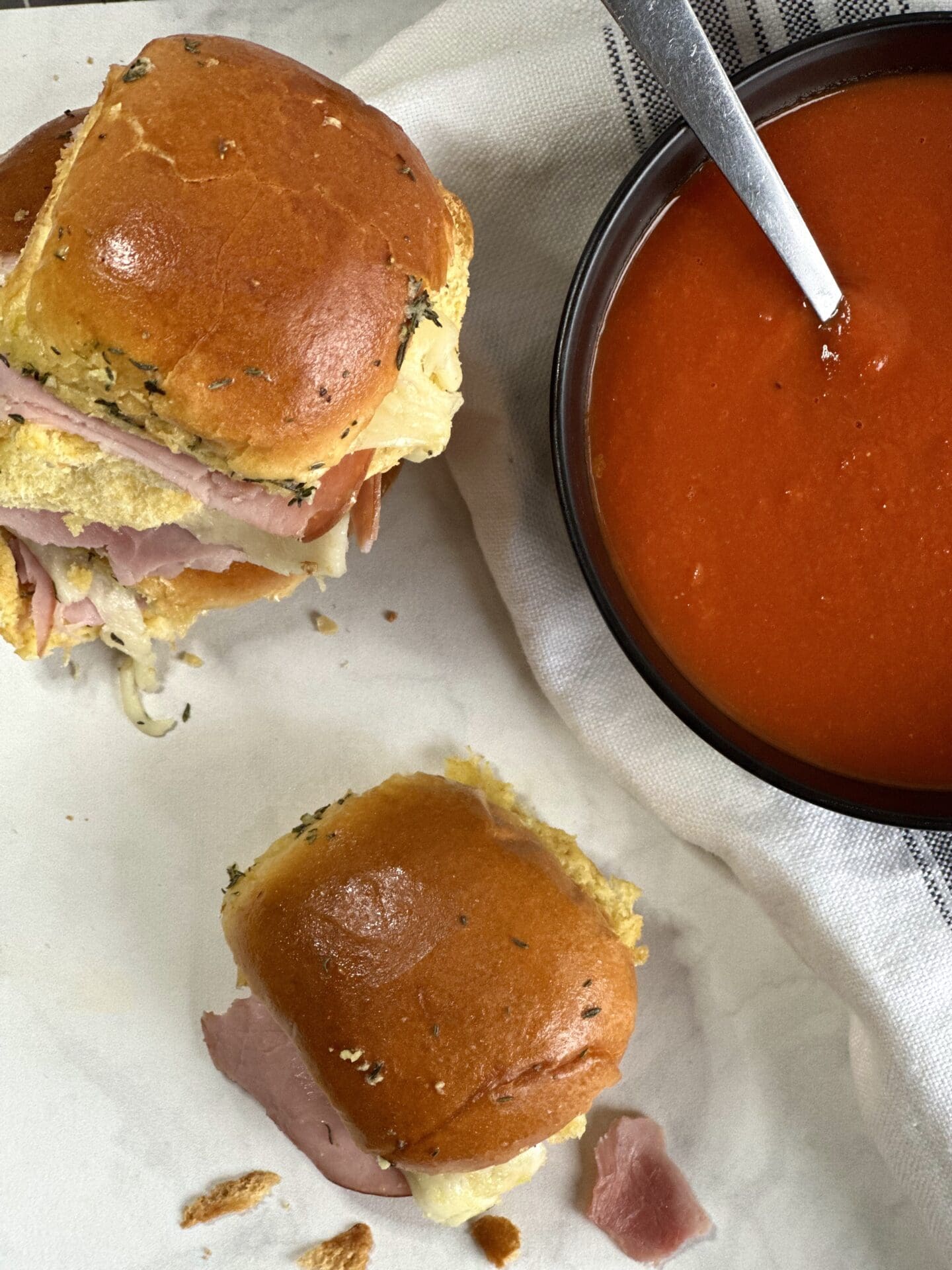 C1D153A9-0C31-41CE-A08B-7C655D09BB12 Hot ham and cheese sliders on golden buns seen from above with a bowl of homemade tomato soup.