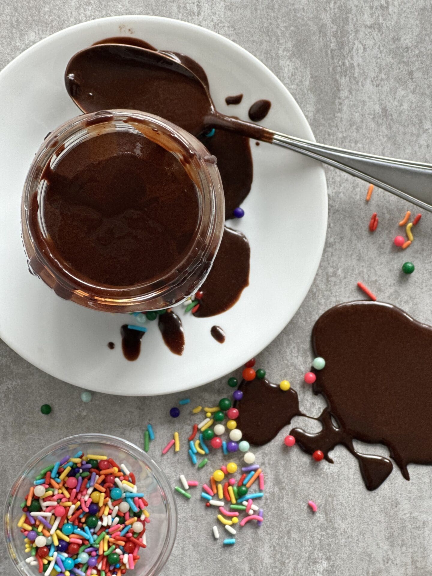 86AF103C-8828-4454-9E4F-6E35C3BC52D6 Jar of Hot Chocolate Fudge Sauce seen from above, surrounded by spilled sauce and colourful sprinkles