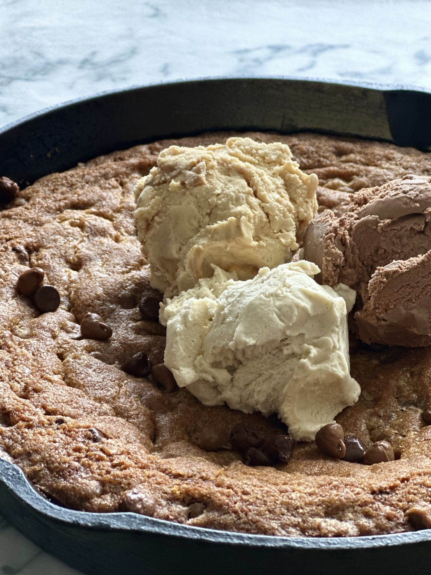 D8A3606E-5231-4DF7-AB12-D80B7A1F7BF3 Cast iron pan full of a giant chocolate chip cookie, topped with scoops of vanilla, chocolate and caramel ice cream