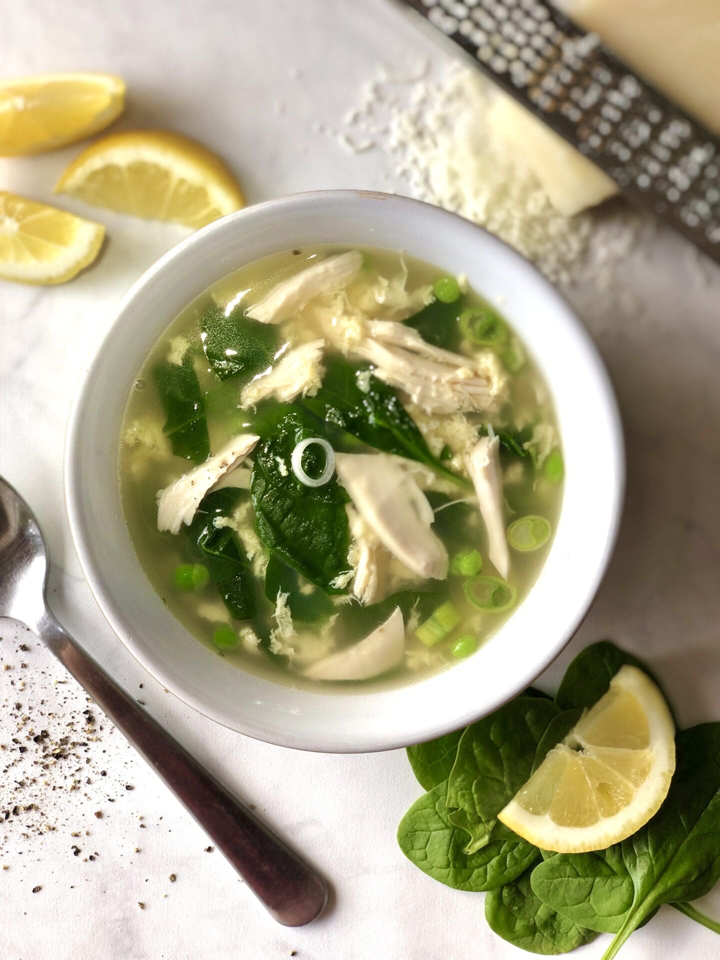 42986BB9-5B0A-4C90-B61A-8415F4B175B7 Bowl of spring straciatella soup with chicken and vegetables, with lemon wedges and freshly grated parmesan cheese.