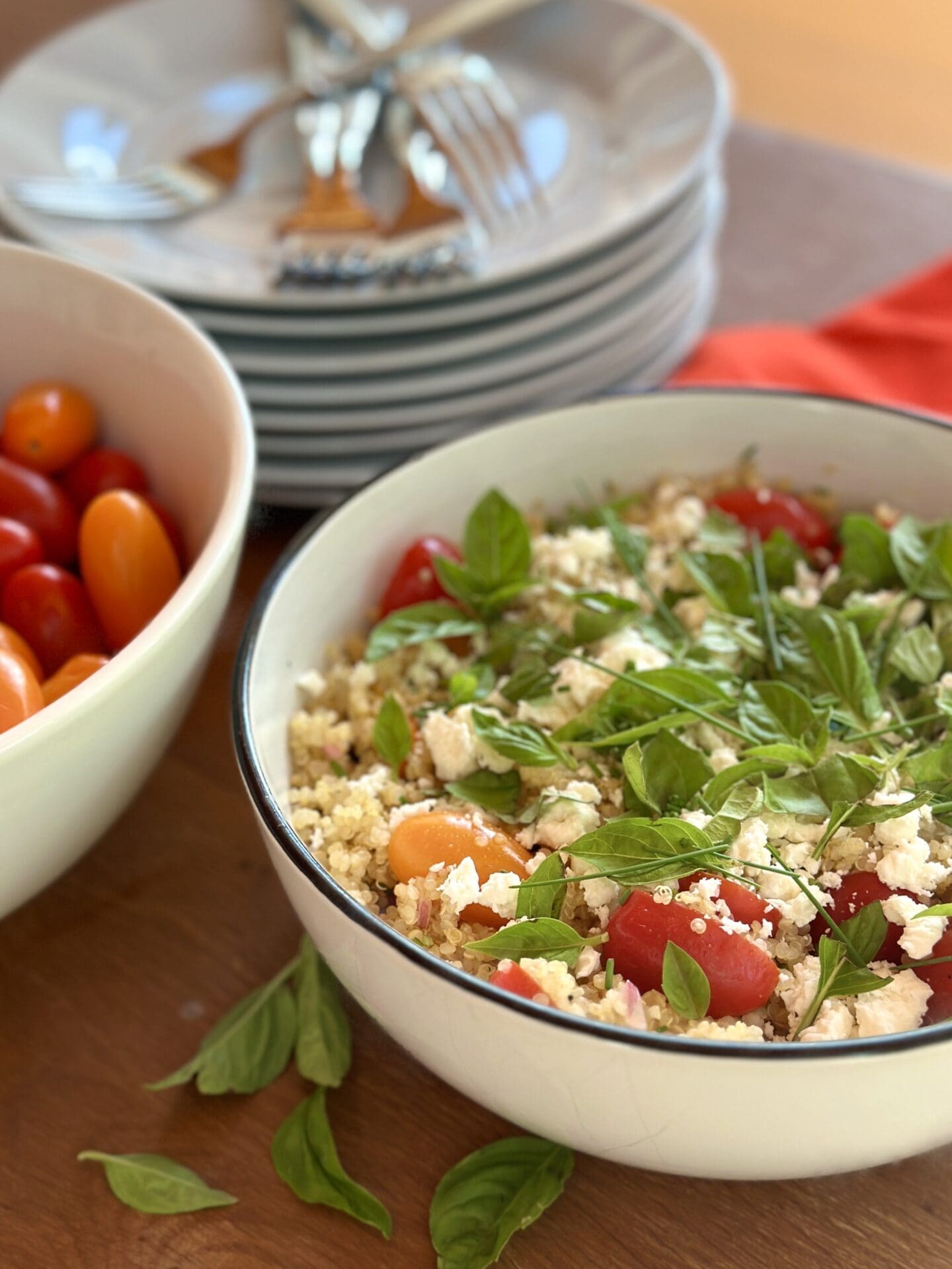 IMG_9067 A large white bowl of cherry tomato quinoa salad garnished with fresh basil is seen, with a stack of white plates and forks in the background