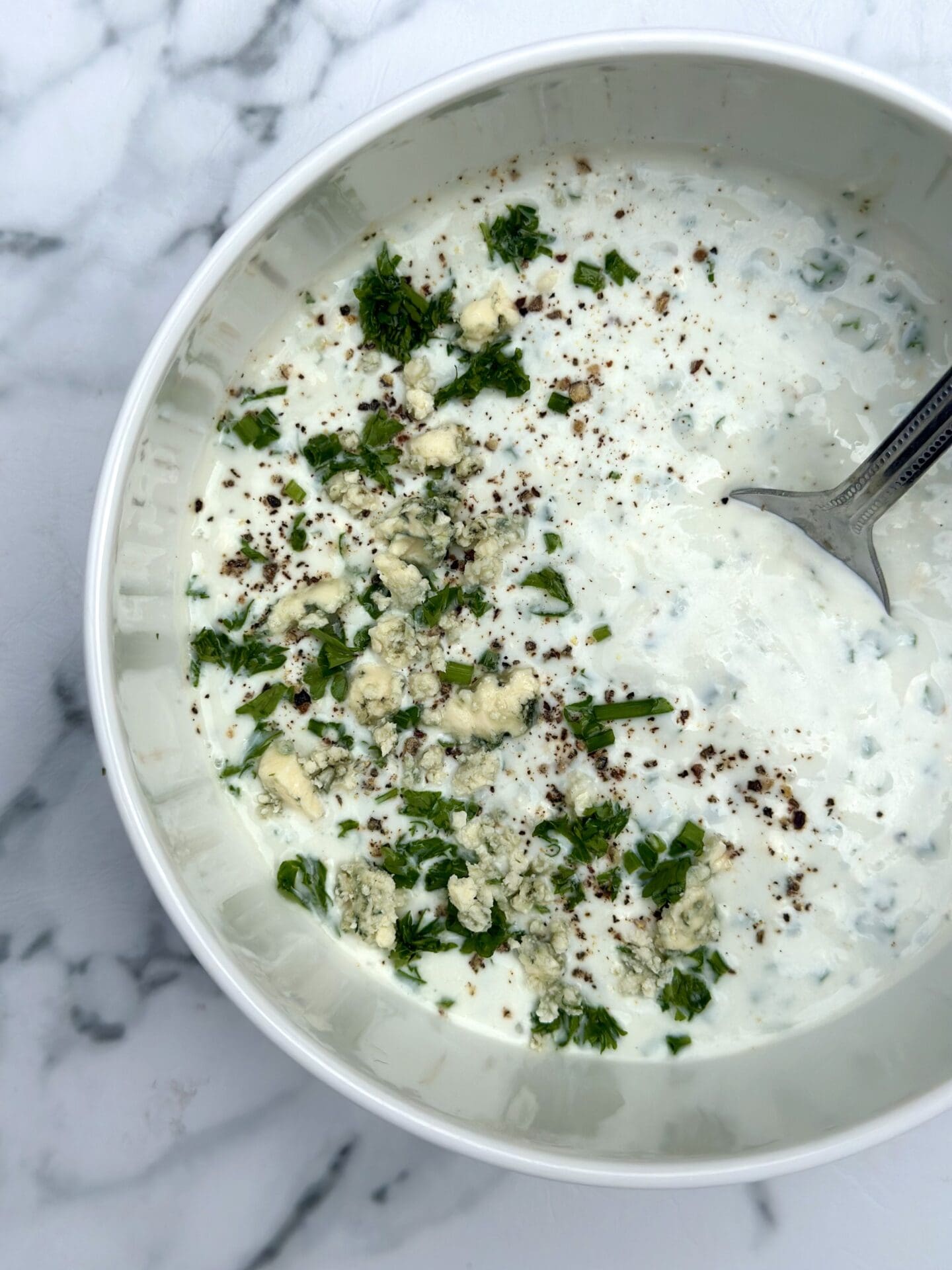 IMG_9317 Bowl of homemade blue cheese dressing seen from above garnished with crumbled blue cheese and fresh herbs, sitting on a white marble table