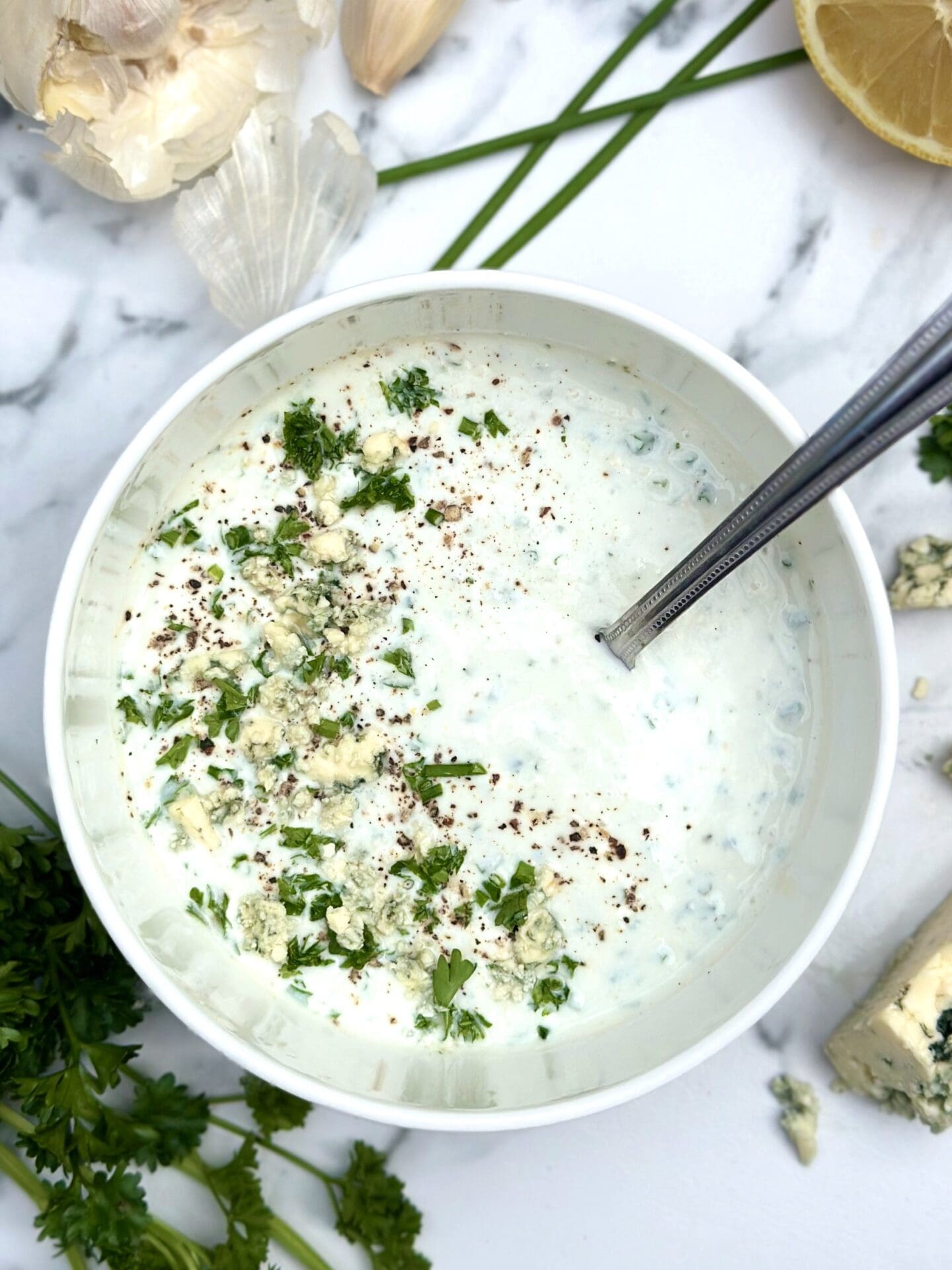 IMG_9325 Bowl of homemade blue cheese dressing seen on a white marble countertop surrounded by lemon, garlic and fresh herbs