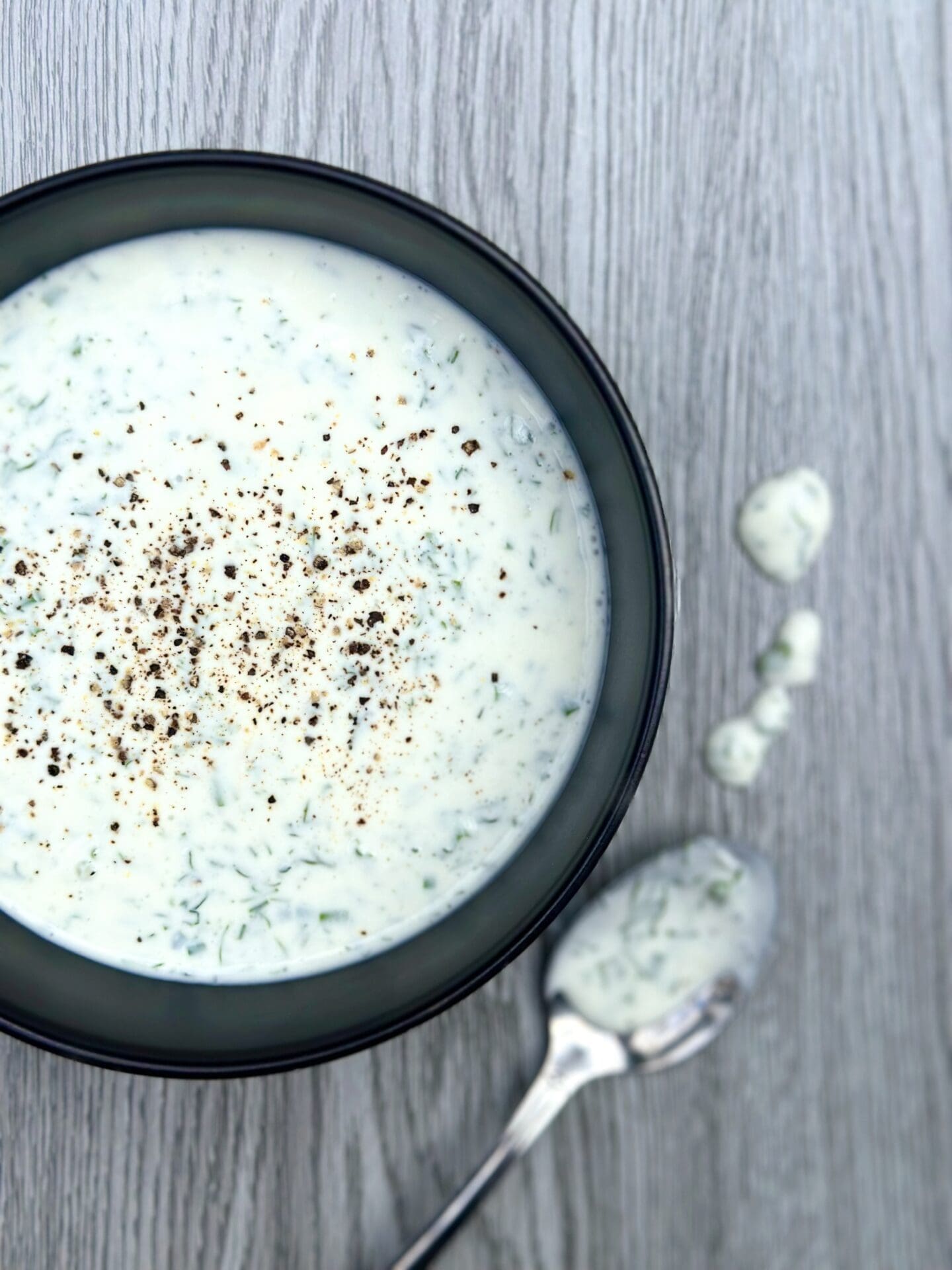 IMG_9331 A bowl of creamy and fresh homemade buttermilk ranch dressing, with a spoon and drips of the dressing on a grey wood board