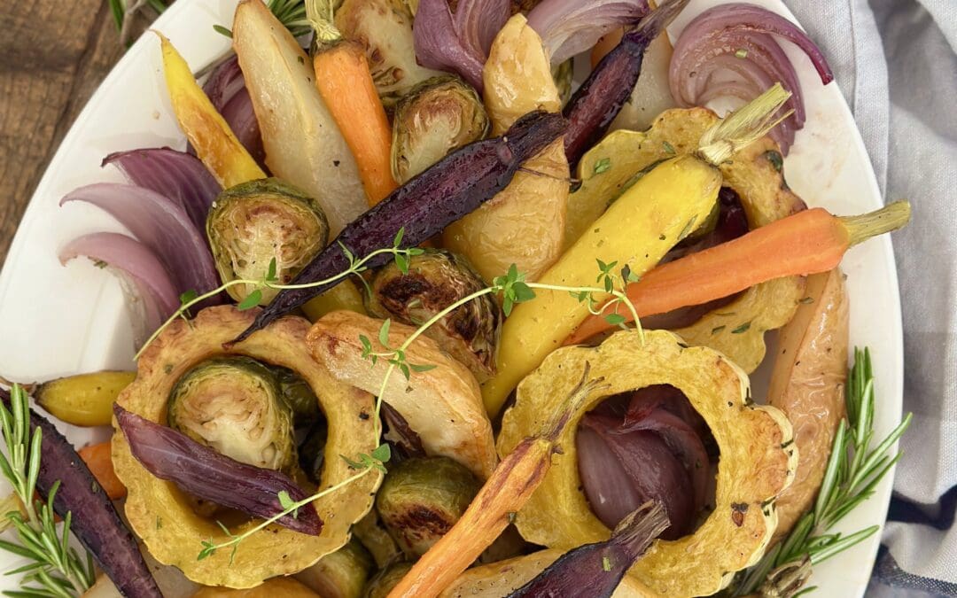 A platter of Maple Butter Roasted Vegetables with Pears is seen from above on a wood table surrounded by fresh rosemary and a grey striped kitchen cloth.