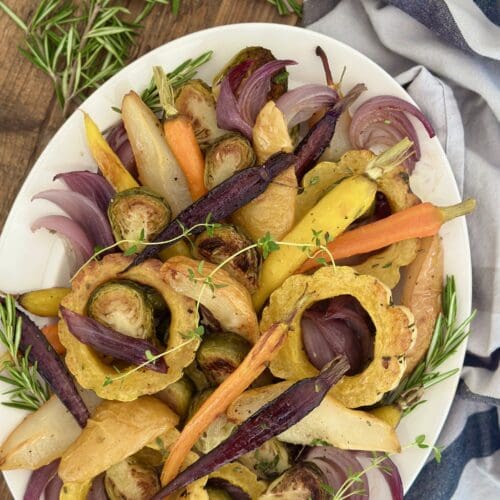 A platter of Maple Butter Roasted Vegetables with Pears is seen from above on a wood table surrounded by fresh rosemary and a grey striped kitchen cloth.