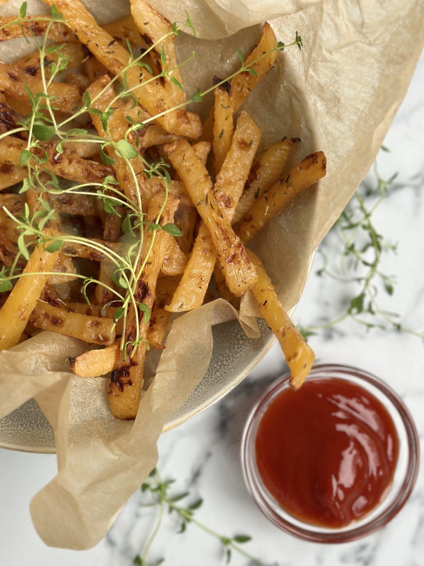 A brown paper lined bowl of golden rutabaga fries garnished with fresh thyme sits on a white marble counter top along with a small bowl of red ketchup