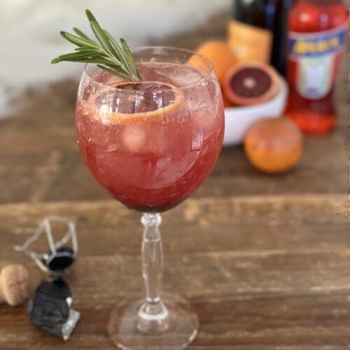 A blood orange and black currant Aperol spritz garnished with rosemary sits on a wood table