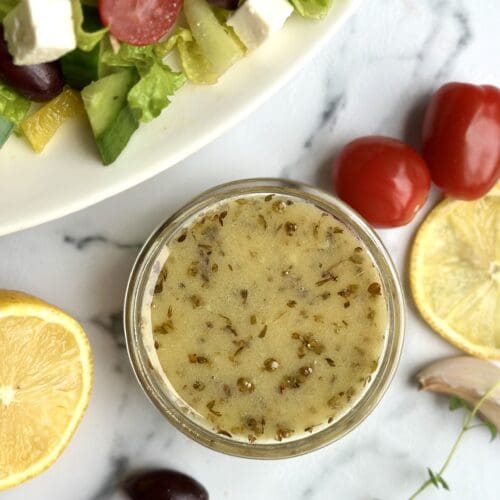 a glass dish of homemade Greek salad dressing is seen from above surrounded by lemons and fresh vegetables
