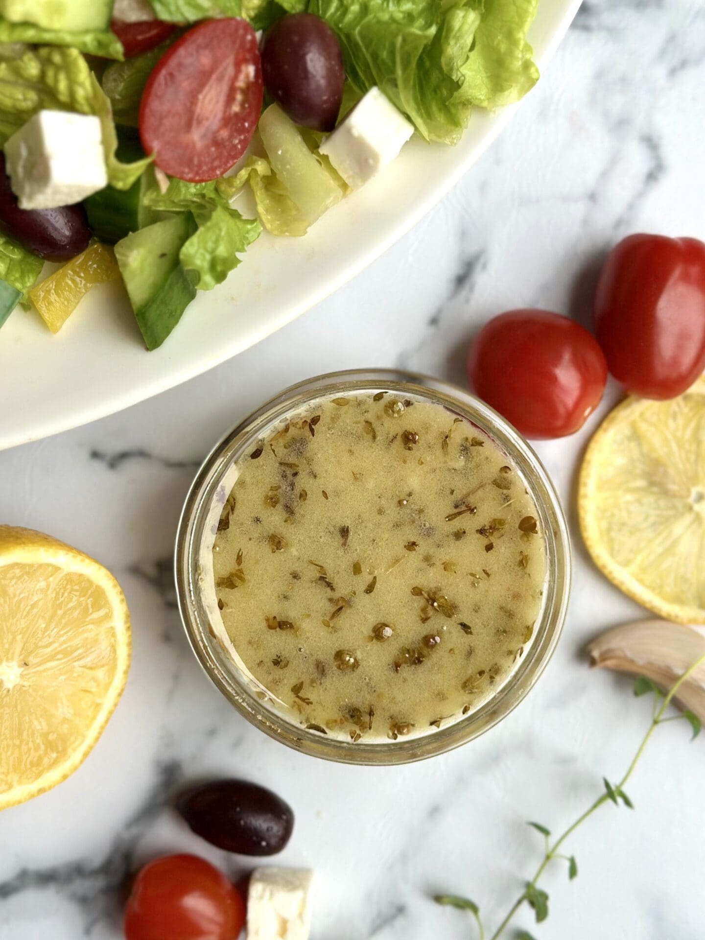 a glass dish of homemade Greek salad dressing is seen from above surrounded by lemons and fresh vegetables