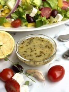 a bowl of Greek salad dressing sits on a white marble counter with a Greek salad seen in the background
