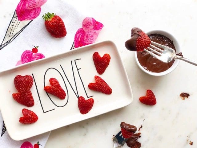 Heart shaped strawberries are seen on a platter with the word Love. A dish of chocolate is nearby for dipping.