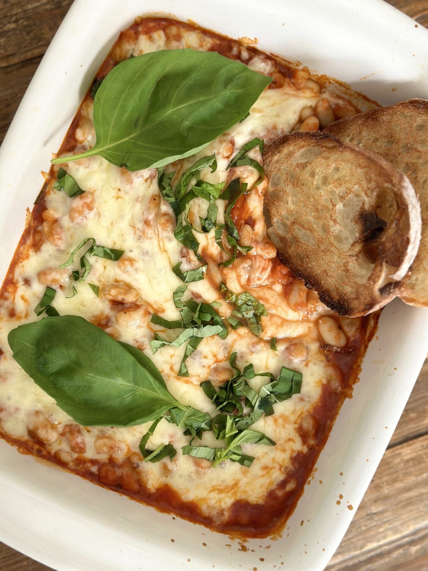 A pan of Easy Cheesy Beans is seen from above, garnished with fresh basil and slices of garlic bread