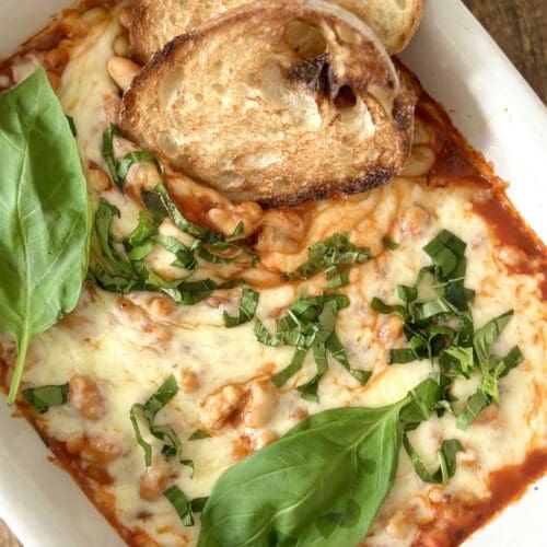 A pan of Easy Cheesy Beans is seen from above garnished with fresh basil and slices of garlic bread