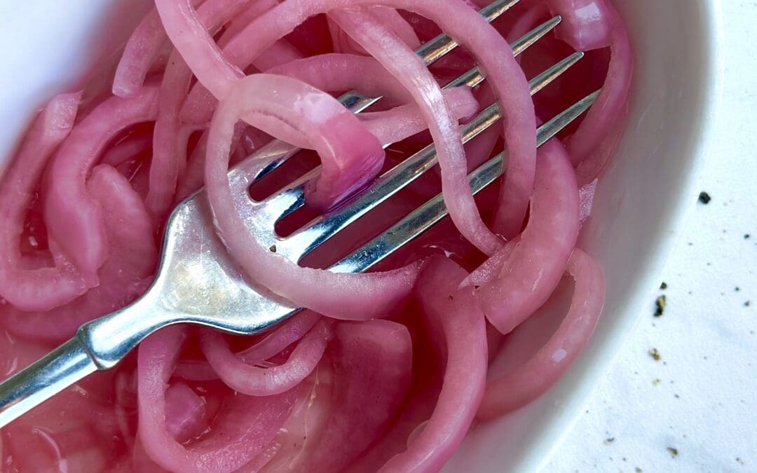 A dish of homemade bright pink pickled red onions tangled in the tines of a fork is seen from above on a white marble counter