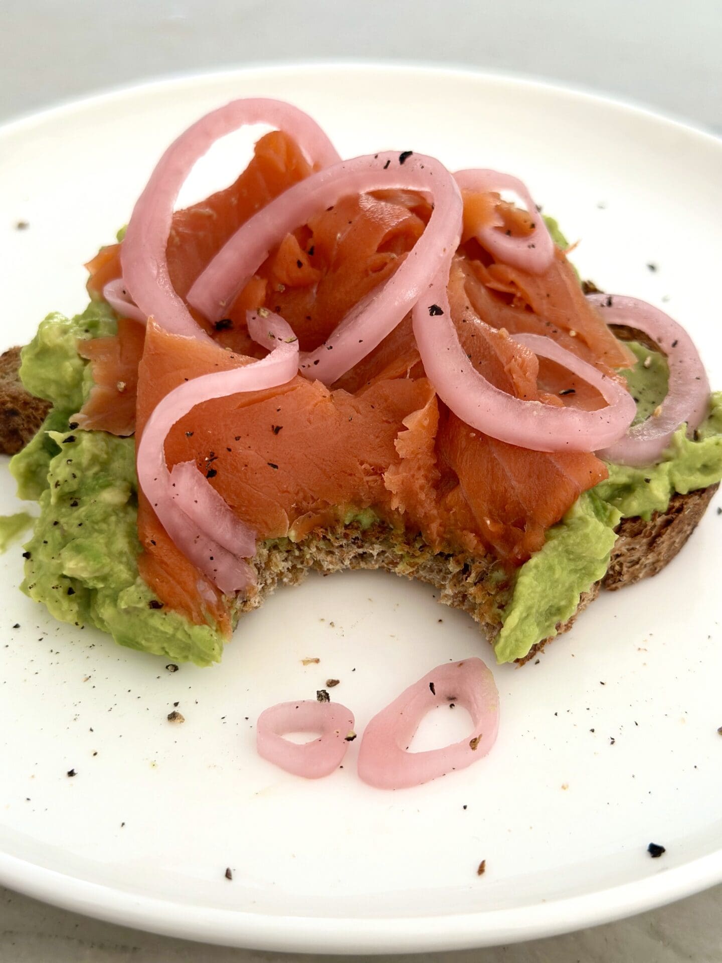 IMG_1296 Avocado toast with smoked salmon and pickled red onions sits on a white plate. A large bite has been taken out of the avocado toast