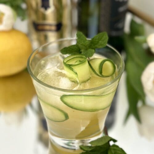 An Irish Maid Cocktail garnished with cucumber curls and fresh mint sits on a sparkling bar surrounded by bottles of Irish Whiskey and St Germain Elderflower Liqueur