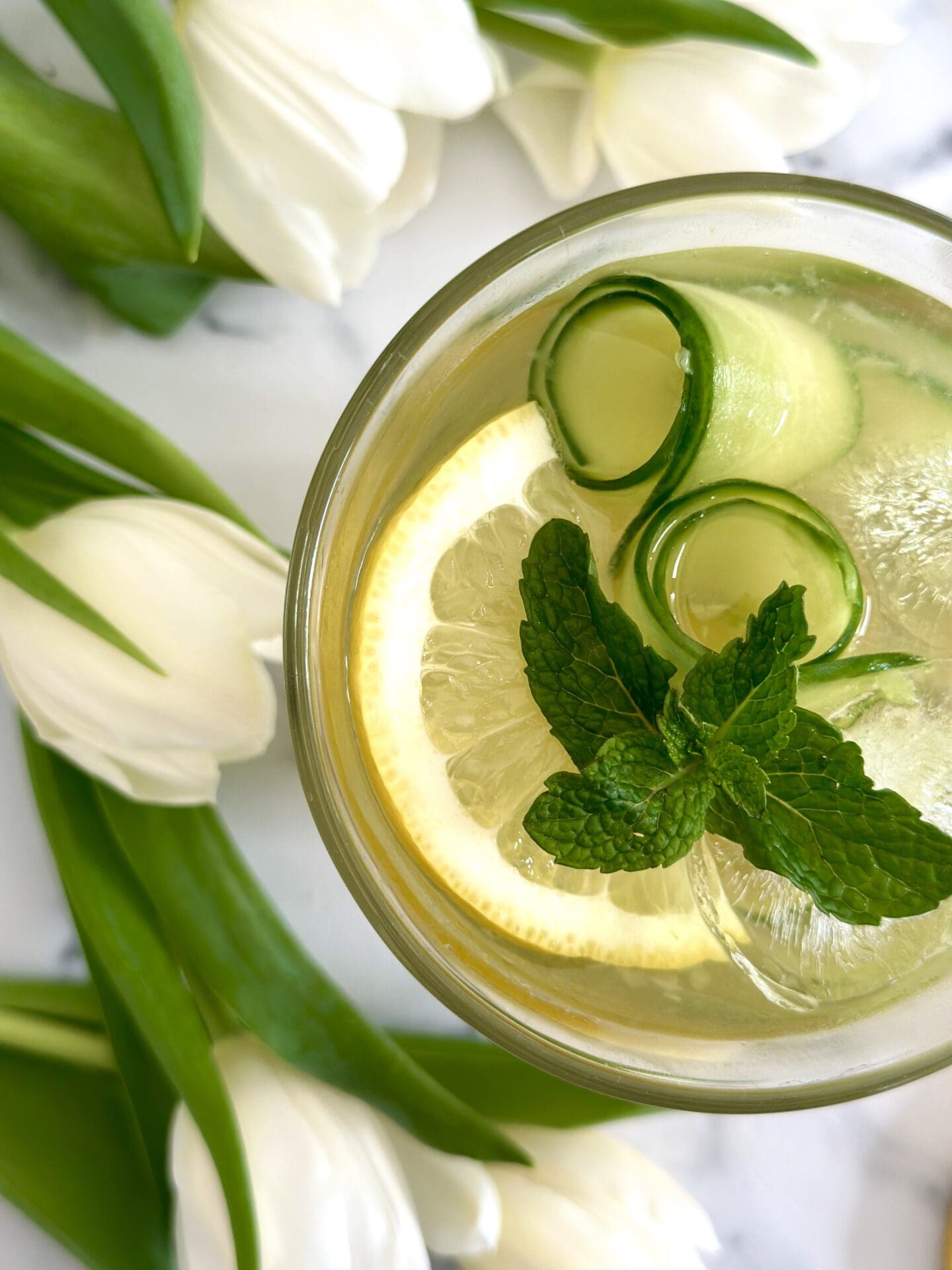 IMG_1357 An Irish Maid Cocktail is seen from above garnished with cucumber curls, lemon slices and fresh mint and surrounded by white tulips