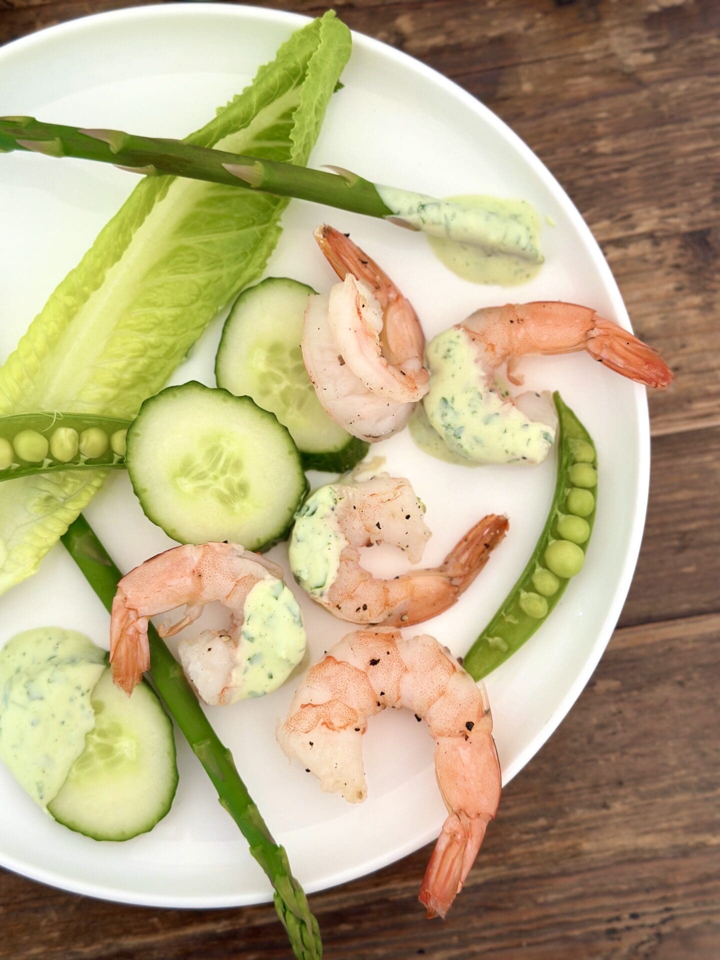 IMG_1470 A white plate with a serving of roasted shrimp and spring vegetables with creamy herb dip is seen from above on a wood table
