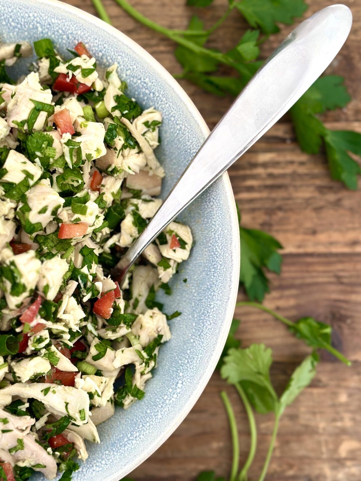 Chimichurri Chicken Salad Half a bowl of chimichurri chicken salad is seen from above on a wood table surrounded by fresh parsley and cilantro