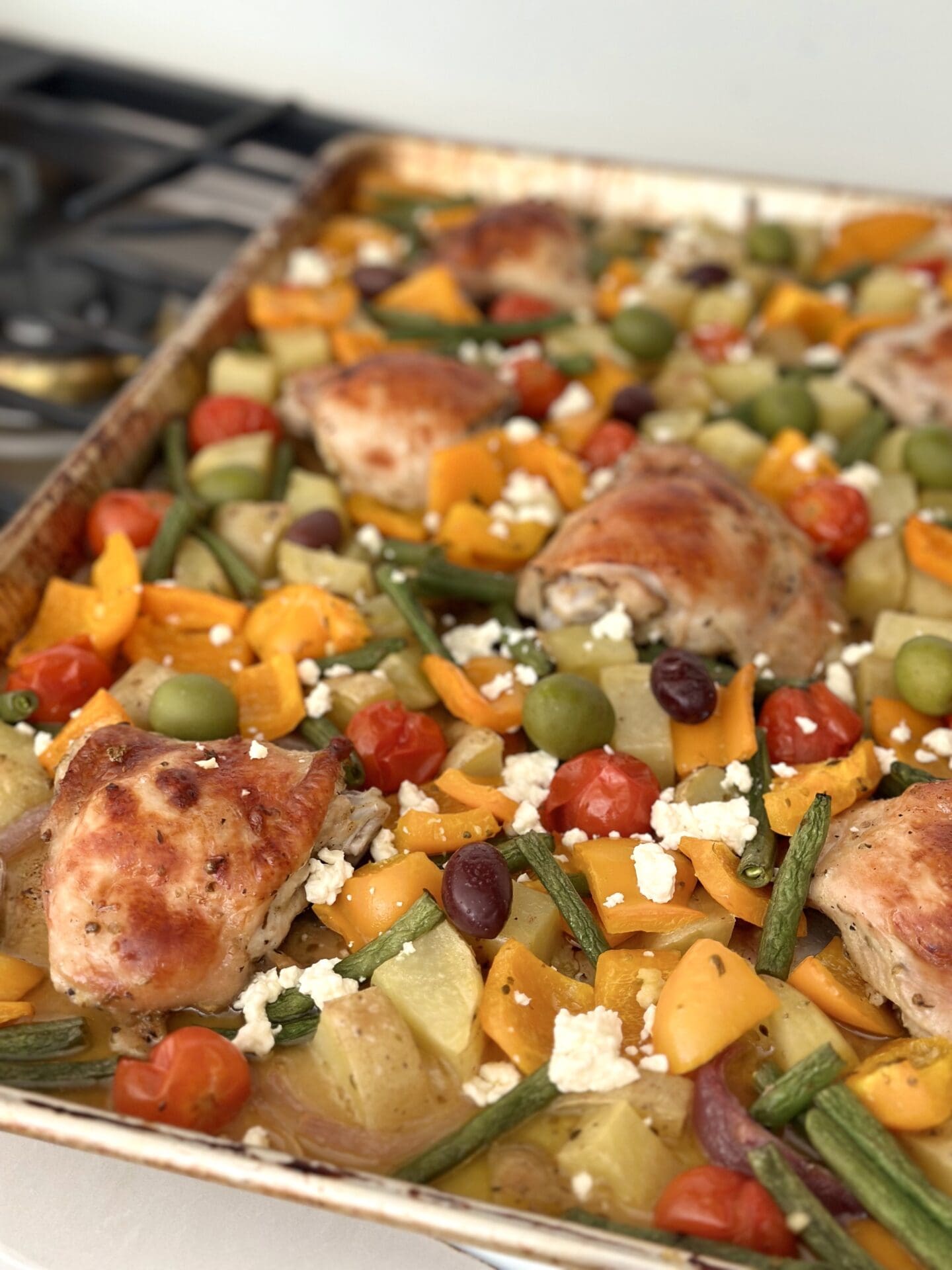 IMG_1683 A large pan of Greek Sheet Pan Dinner that has roasted to perfection in the oven is seen resting on a cook top. The dinner features golden chicken thighs nestled amongst roasted orange bell peppers, chopped potatoes, red onion wedges and green beans. The meal is garnished with crumbled feta cheese and olives.