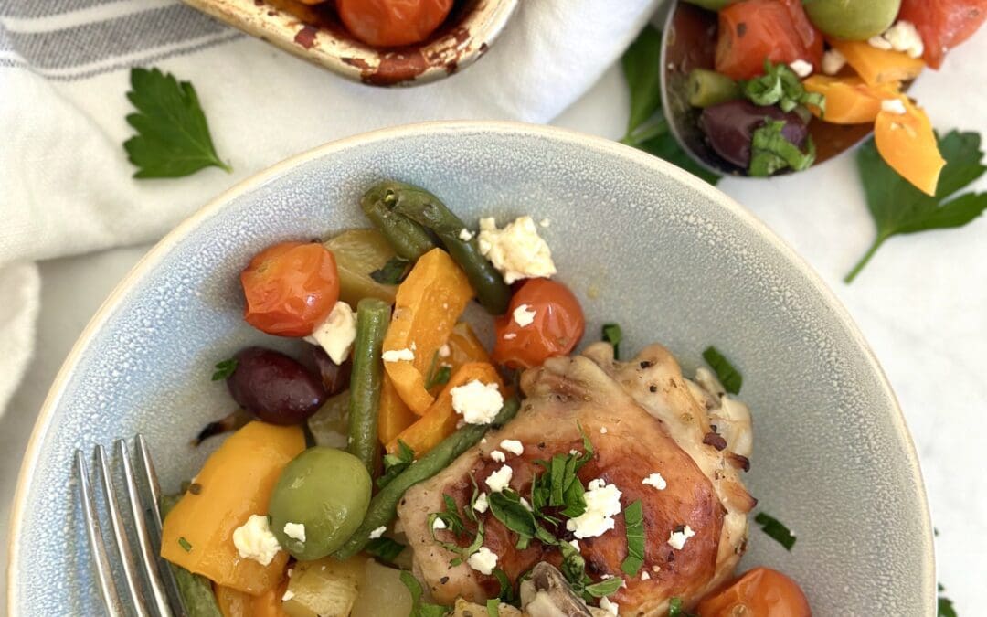 A bowl of sheet pan Greek Chicken dinner is seen from above with golden chicken thighs, orange bell peppers, green beans and potatoes. The dish is garnished with crumbled feta cheese and fresh herbs. The sheet pan and a large spoon with more of the meal are seen in the background.