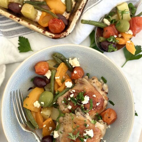 A bowl of sheet pan Greek Chicken dinner is seen from above with golden chicken thighs, orange bell peppers, green beans and potatoes. The dish is garnished with crumbled feta cheese and fresh herbs. The sheet pan and a large spoon with more of the meal are seen in the background.