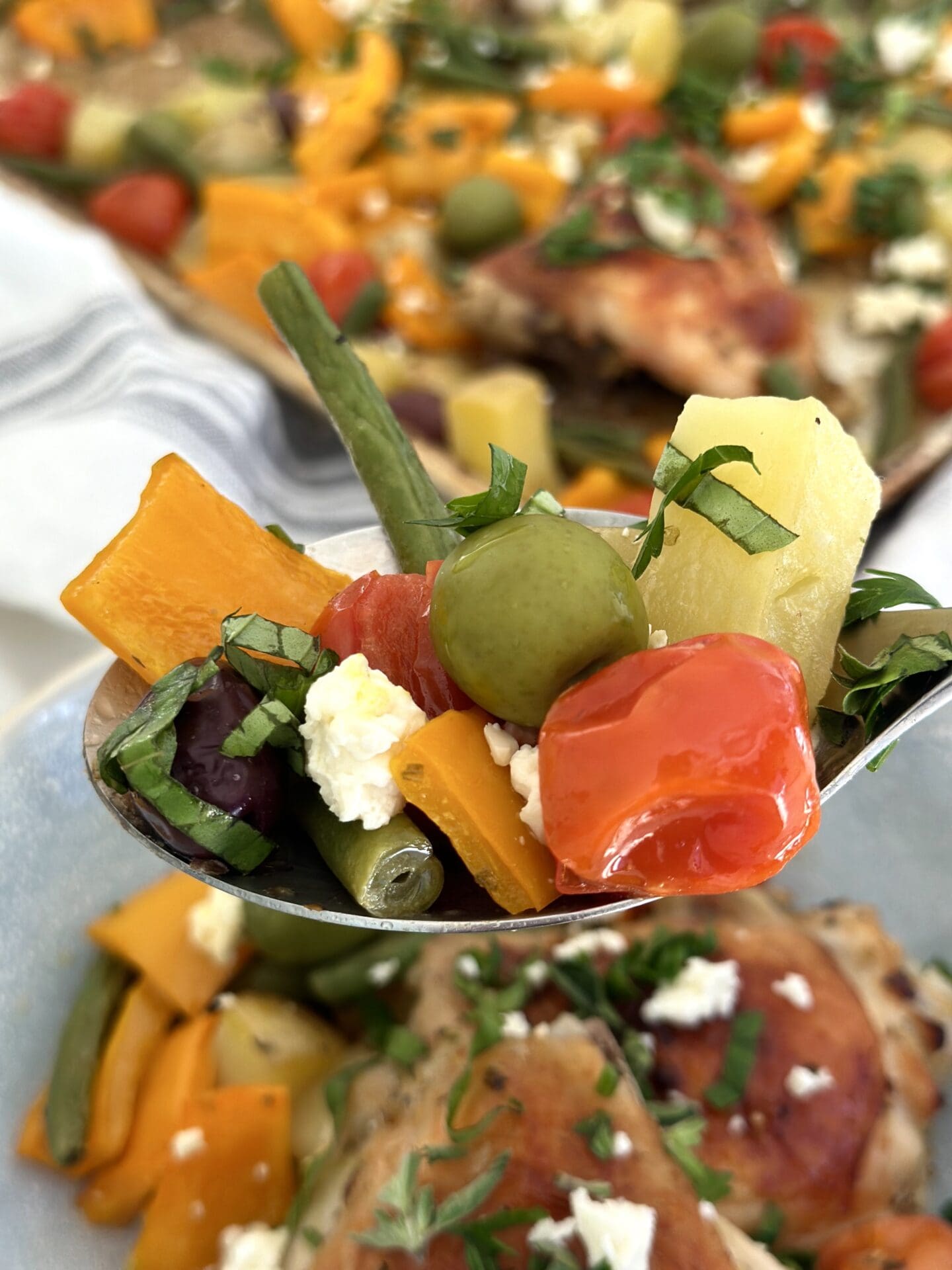 IMG_1711 A large spoonful of vegetables, olives, feta cheese and fresh herbs is seen in the foreground with Greek Chicken Sheet Pan Dinner seen in behind
