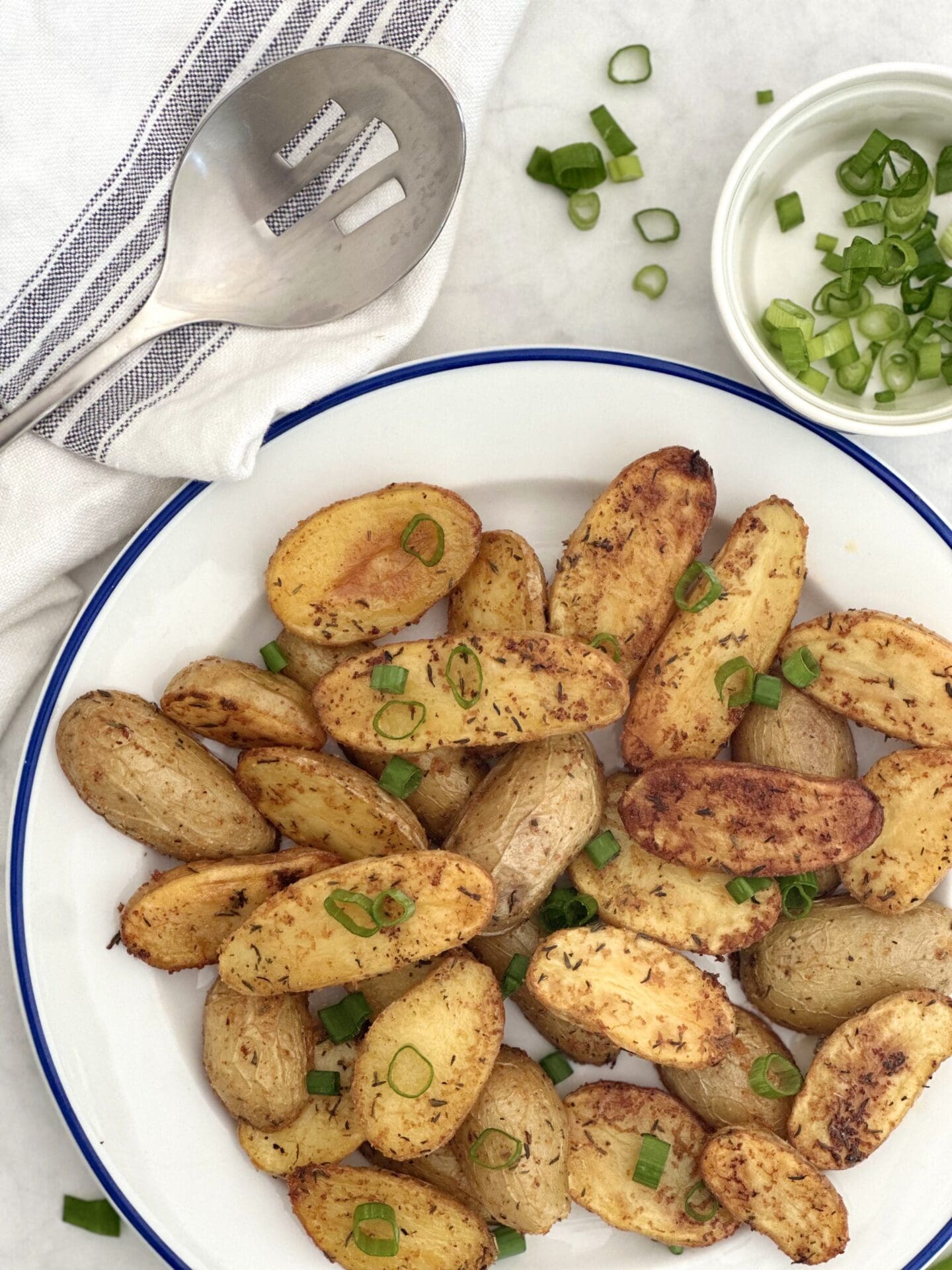 IMG_2040 A blue trimmed plate of golden and crispy roasted fingerling potatoes is seen from above with a serving spoon and chopped green onion for garnishing