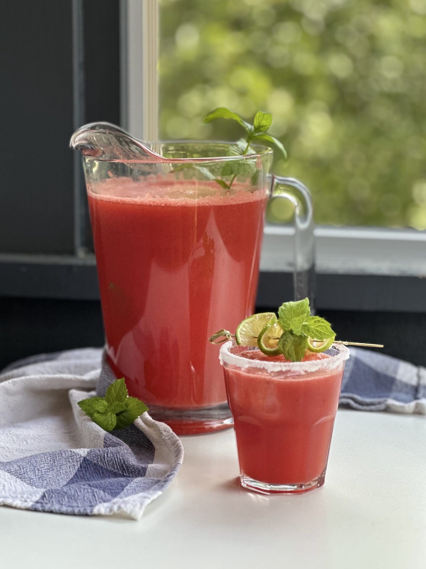 IMG_2379 A large pitcher of freshly made watermelon agua fresca sits by a window, with a sugar rimmed glass of the beverage, garnished with lime and mint, sits in the foreground.