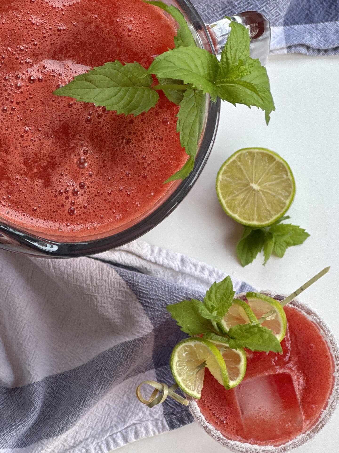 IMG_2383 A glass and a pitcher of freshly made watermelon agua fresca are seen from above, surrounded by a blue checkered cloth and fresh mint and lime