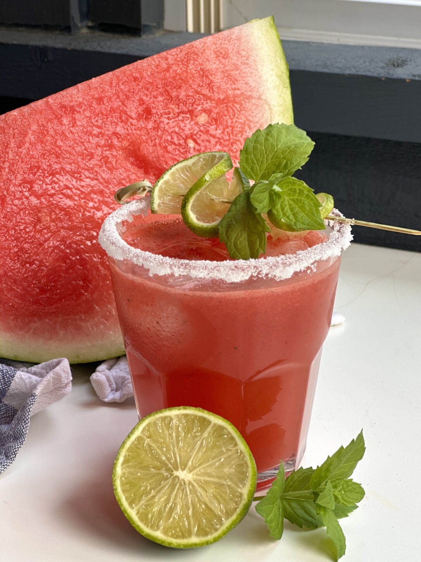 A sugar rimmed glass of freshly made watermelon agua fresca sits on a table surrounded by fresh mint, lime and a large slice of watermelon