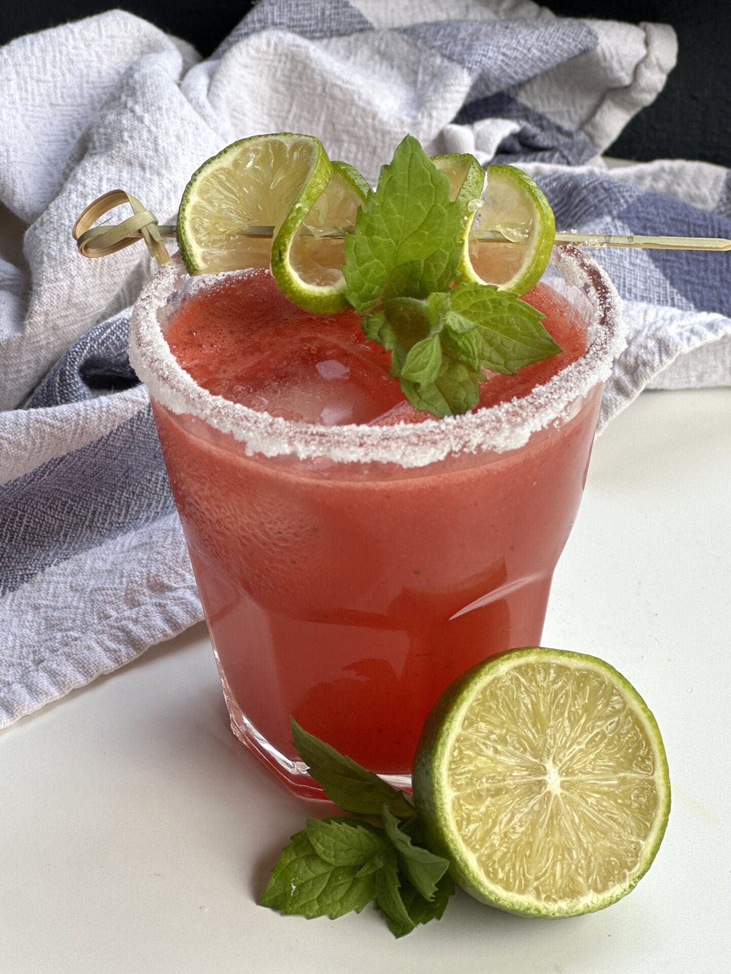 IMG_2402 A sugar rimmed glass of watermelon agua fresca garnished with fresh mint and lime sits on a white table.