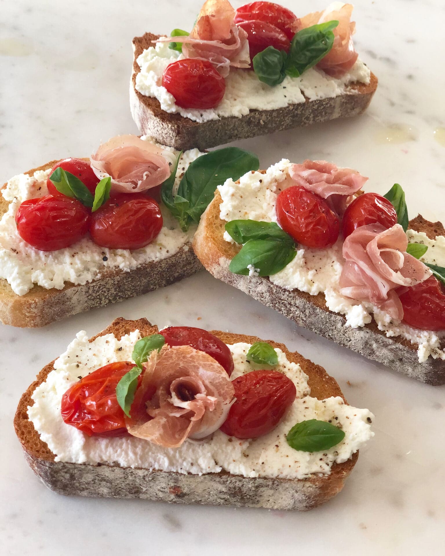 FA26C601-027A-4CC9-9CE4-0D09B216B5FB Thick slices of toast are seen slathered in homemade ricotta cheese and topped with sliced cherry tomatoes, fresh basil and prosciutto rosettes
