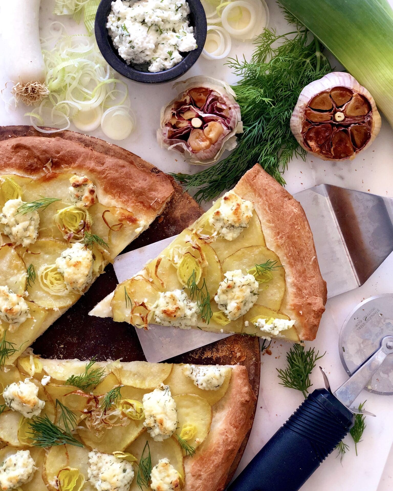 IMG_1523 A potato, leek and ricotta pizza is seen from above, surrounded by heads of roasted garlic and fresh herbs