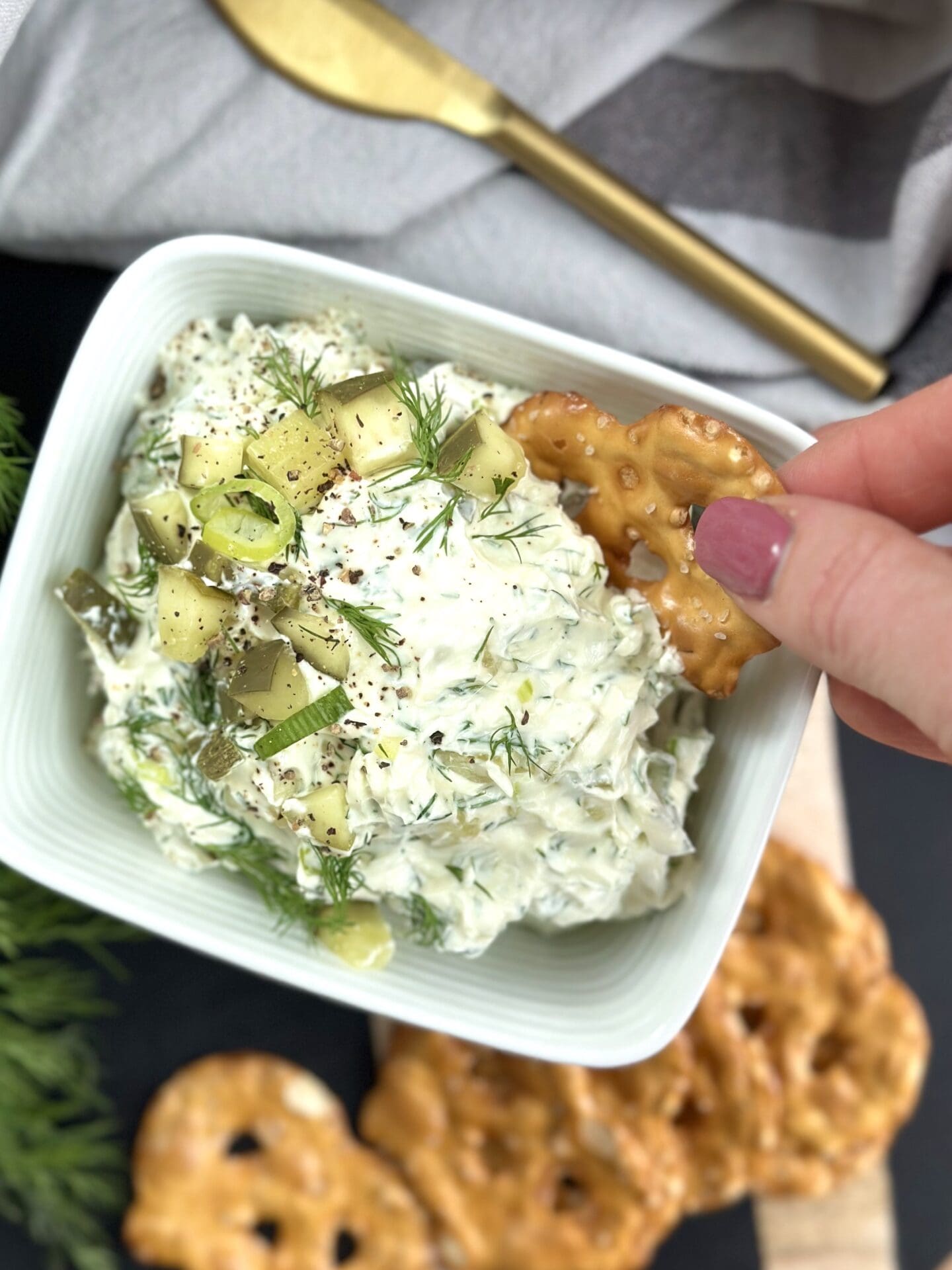 IMG_3295 A woman's hand is seen dipping a pretzel chip into a bowl of dill pickle dip garnished with chopped dill pickles and fresh dill.