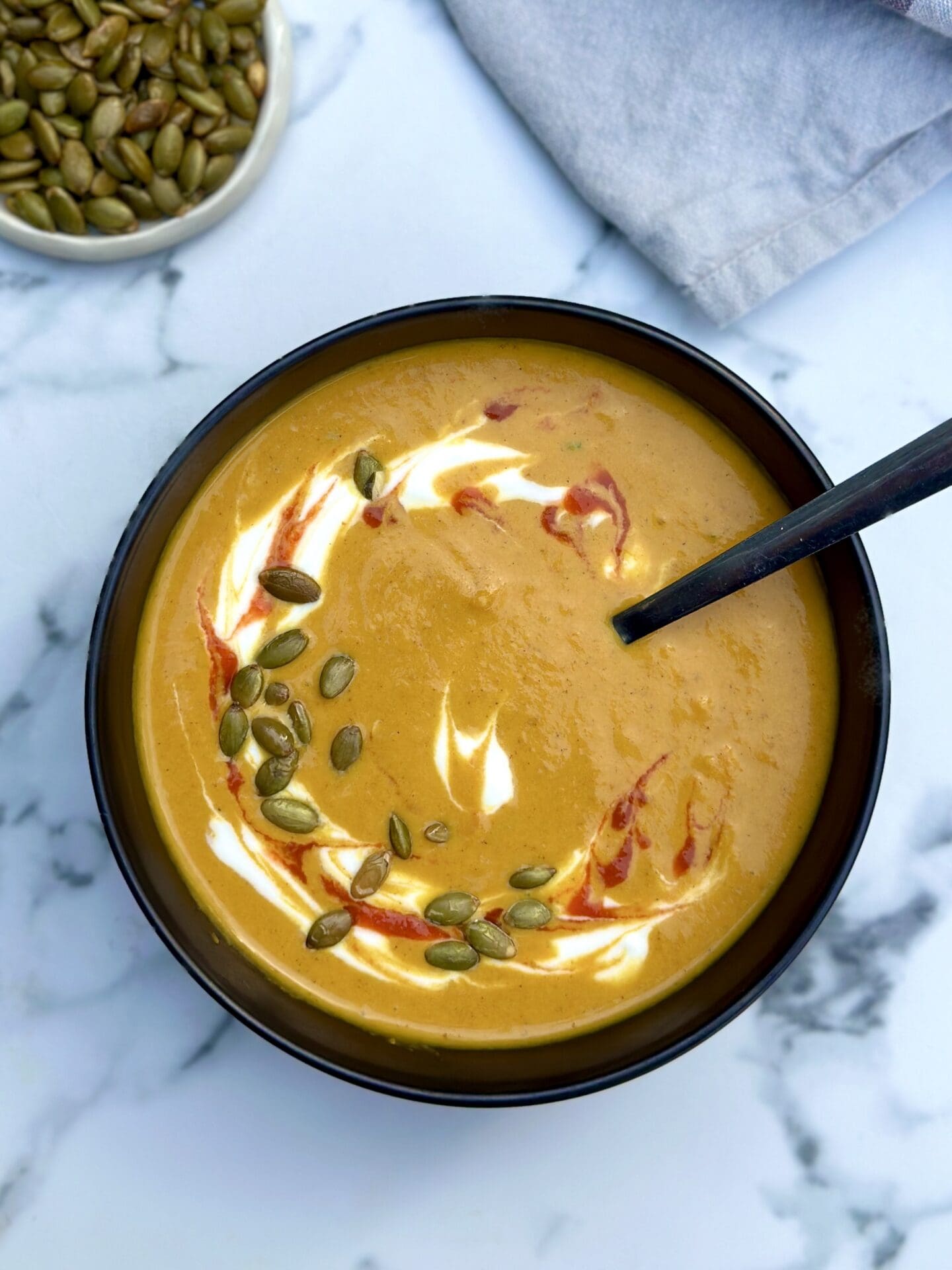 IMG_9794 A black bowl filled with golden creamy pumpkin soup is seen from above garnished with swirls of yogurt and hot sauce and sprinkled with green pumpkin seeds