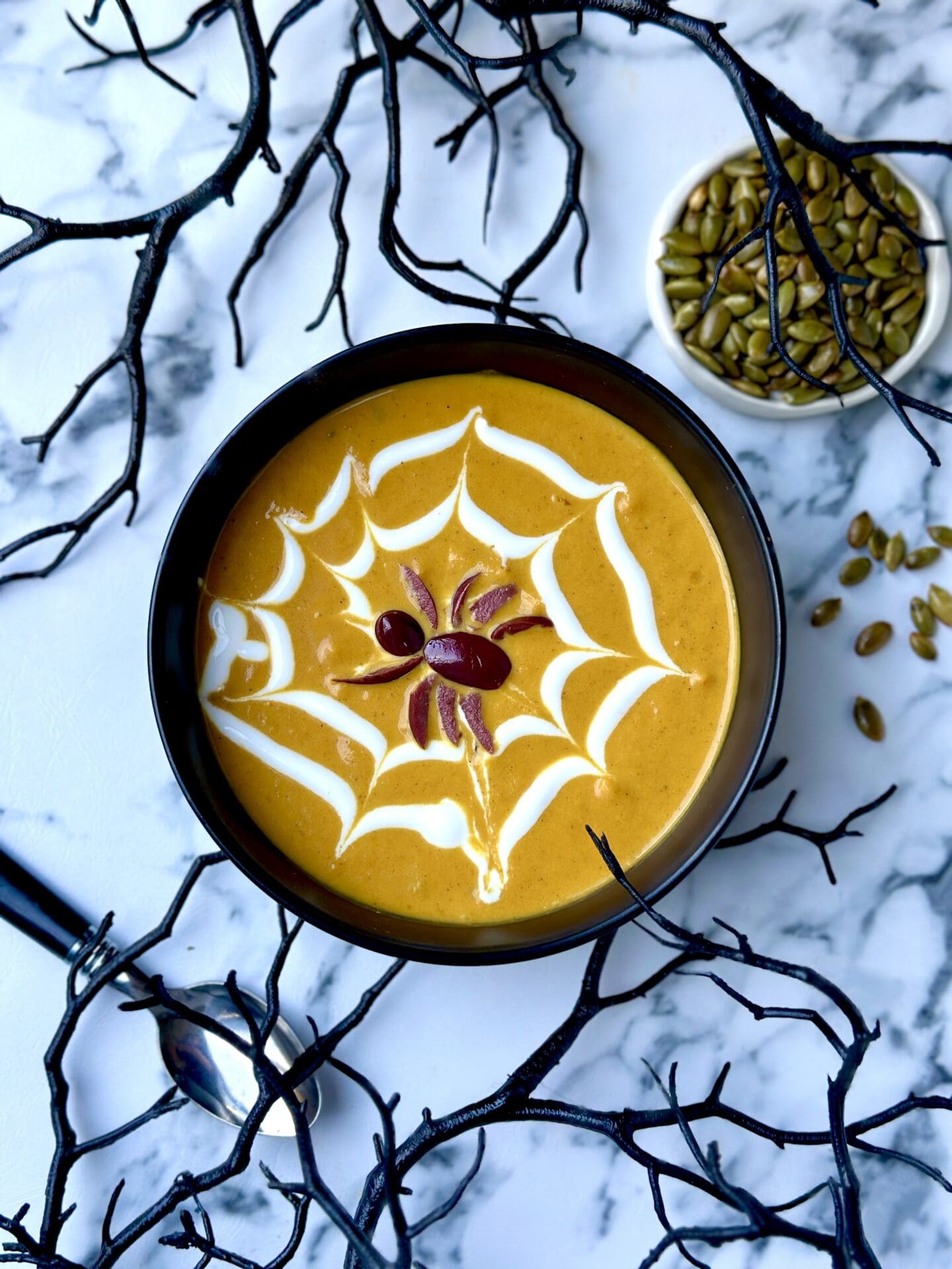 IMG_9800 A black bowl of golden Creamy Pumpkin Soup is decked out for Halloween with a yogurt web and black olive spider garnish. The bowl rests on a black veined white marble table and is surrounded by black branches for a spooky effect.