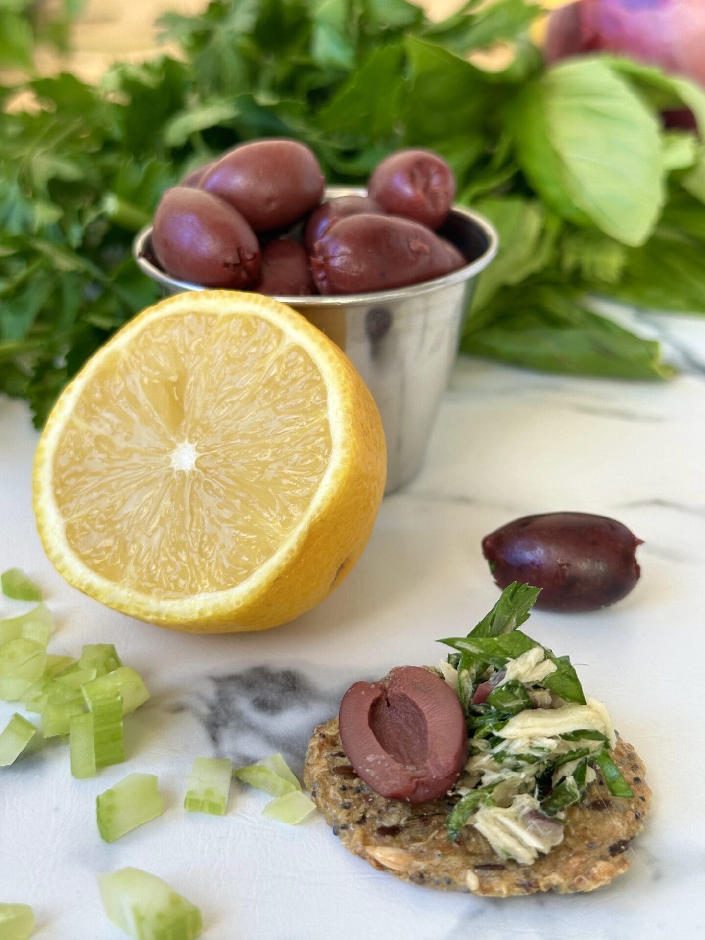 IMG_3491 A cracker topped with Italian tuna salad sits on a white marble table, surrounded by lemons, black olives, chopped celery and lots of fresh green herbs.