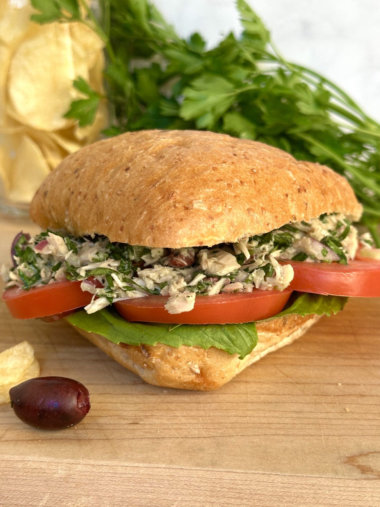 IMG_3502 A large ciabatta bun is stuffed full of Italian tuna salad, thick red tomato slices and fresh basil leaves. The sandwich sits on a wood serving board, surrounded by black olives, potato chips and parsley.