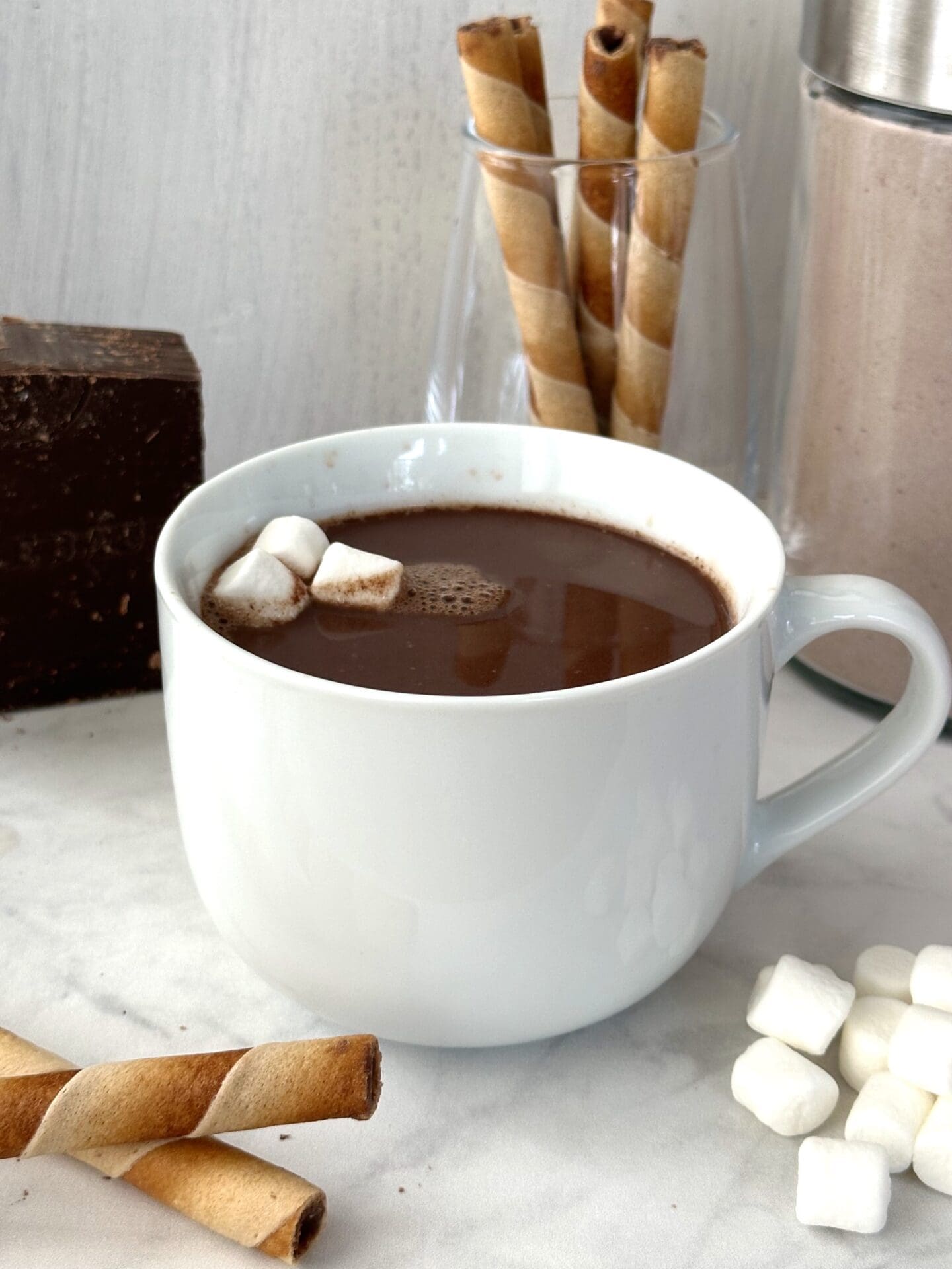 IMG_3610 A large mug of rich dark brown hot chocolate is garnished with mini marshmallows and surrounded by cookie rolls and a big block of dark chocolate