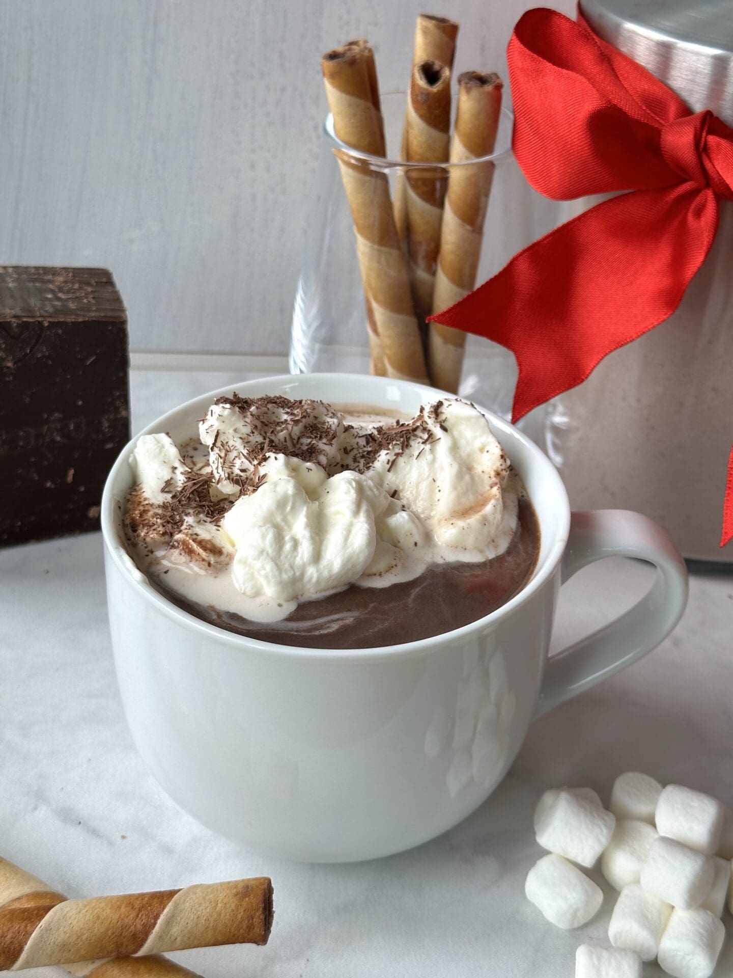 IMG_3615 A large mug filled with whipped cream-topped hot chocolate is surrounded by cookie rolls, marshmallows and a big block of chocolate. A jar of the hot chocolate mix, tied up with a festive red bow for gifting is seen in the background.