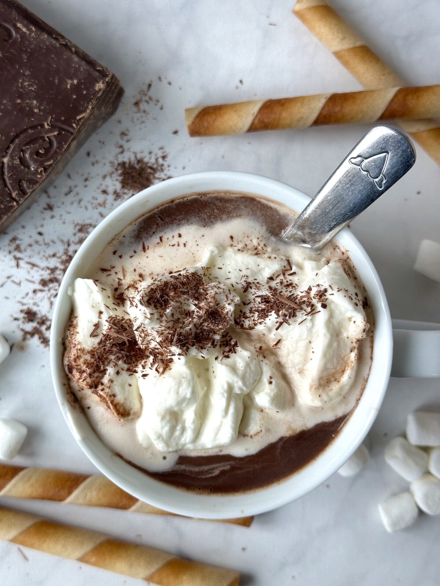 IMG_3622 A big white mug of hot chocolate is seen from above, topped with whipped cream and chocolate shavings. Cookie rolls, a big block of chocolate and piles of mini marshmallows are seen in the background.